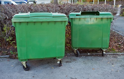 Two large green wheeled waste bins made of durable plastic are positioned side by side on a paved surface near a hedge with brown and green foliage. The bin on the left is slightly larger and has a flat lid with a handle, while the smaller bin on the right features a slightly domed lid with a handle and appears to have a small black wheel visible beneath it. Both bins are set against a background of shrubs and a fence, suggesting an outdoor area possibly in a suburban or urban environment in or around Ruislip. The scene is illuminated by natural daylight, providing clear visibility of the textured, slightly reflective surface of the plastic and the muted tones of the surrounding environment, typical of waste management areas serviced by companies like Office Clearance Ruislip. This image depicts waste collection equipment used for rubbish removal services within local communities in the Ruislip postcode area.