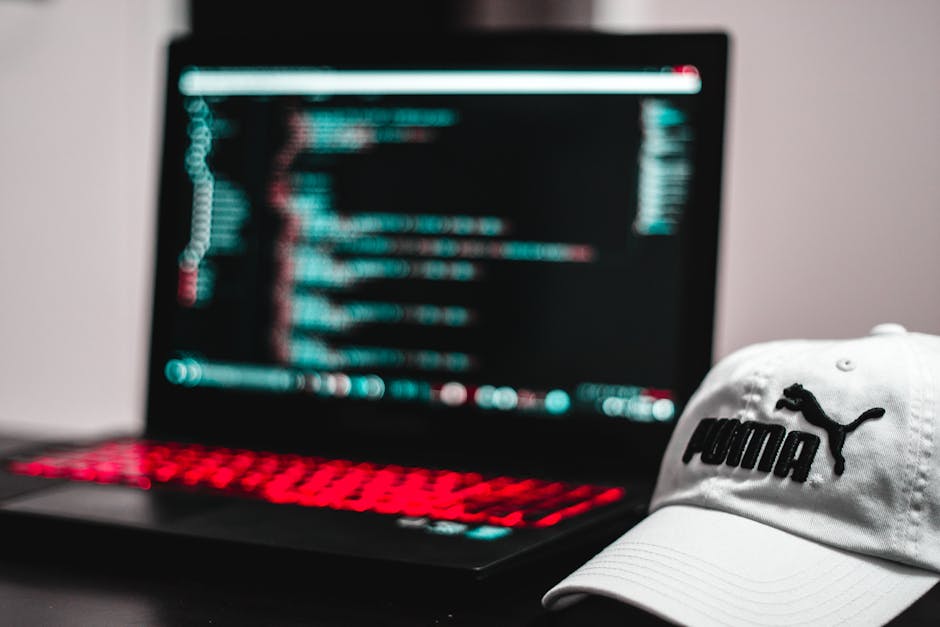 Close-up of a laptop computer with a black casing and a red backlit keyboard, displaying lines of blurred code on a dark screen, positioned on a smooth, dark surface. To the right of the laptop, there