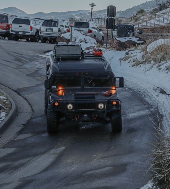 A black utility or military-style vehicle driving along a narrow, curved asphalt road in a snowy environment during daytime. The vehicle has a boxy shape with a textured matte finish, and features two