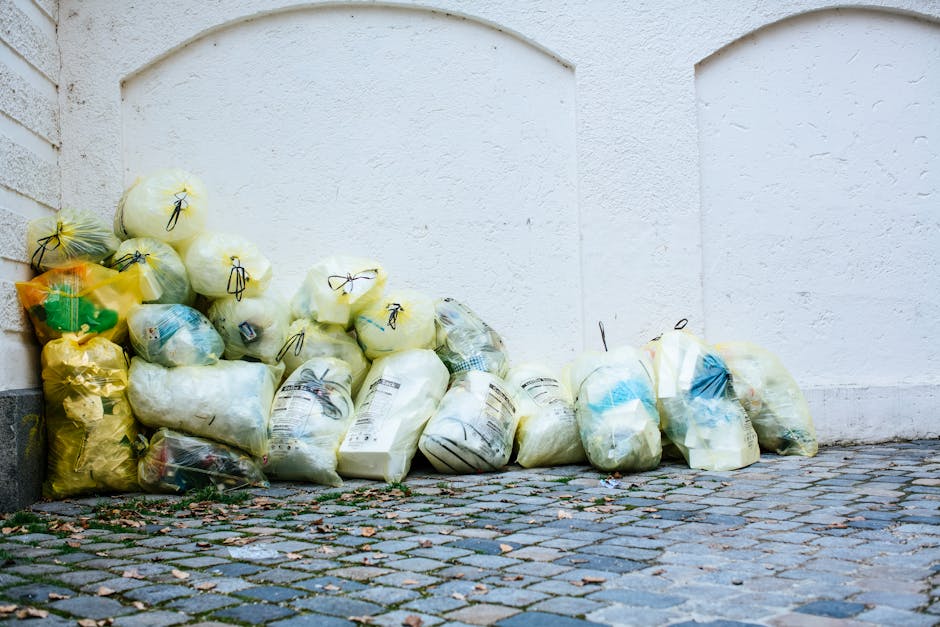 A collection of black rubbish bags and discarded cardboard and paper waste piled in front of a red metal door embedded in a beige, slightly textured wall. The bags are stacked unevenly on a pavement o