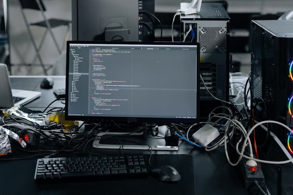 A cluttered desk in a workspace or server room featuring a central flat-screen monitor displaying lines of computer code with syntax highlighting. The monitor is surrounded by various tangled cables a